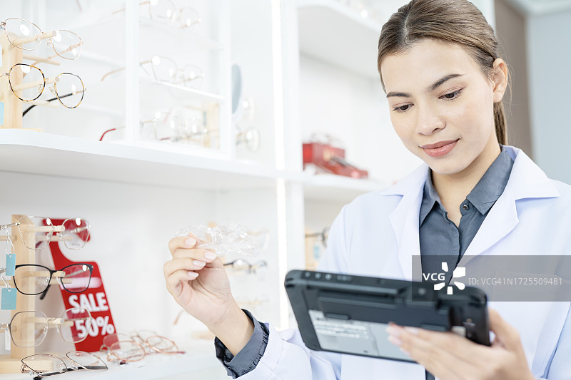 眼睛护理和视力服务。一位女验光师或店主在眼镜店的展示架旁，使用数位平板电脑检查镜框的质量。图片素材