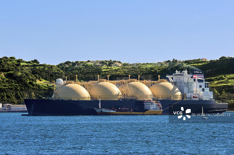 停靠在工业码头旁的液化天然气运输船和阳光明媚的绿色山丘图片素材