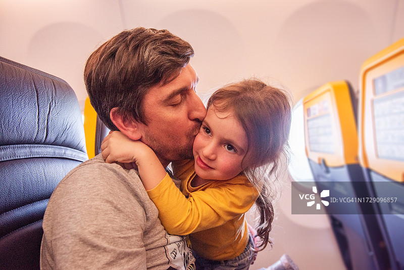 年轻的父亲在飞机舷窗旁亲吻他淘气的小女儿的脸颊图片素材