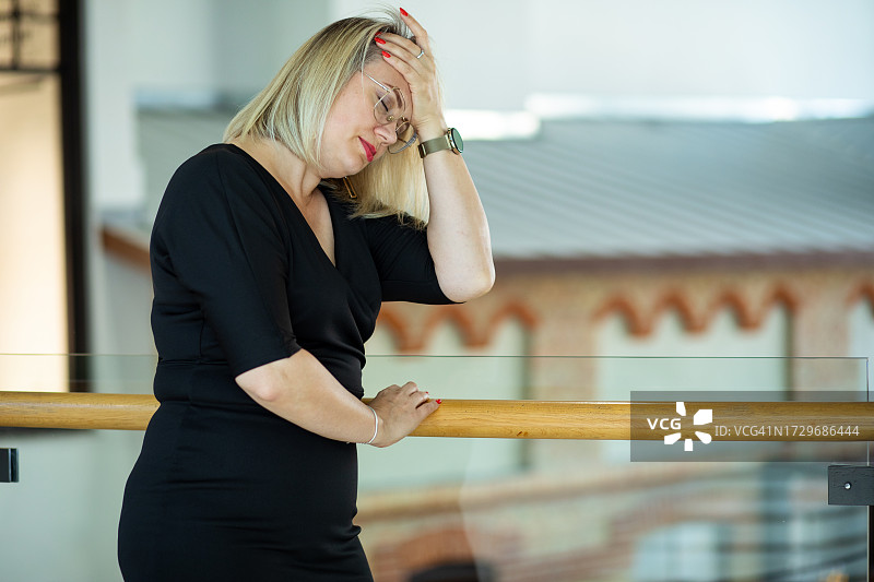 迷人的金发女郎身穿时尚的黑色连衣裙站在栏杆旁，一只手轻轻地触摸着头部图片素材