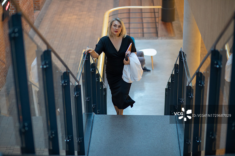 自信的女公司律师，大步走上楼梯，携带笔记本电脑和智能手机，白色西装搭在手臂上图片素材