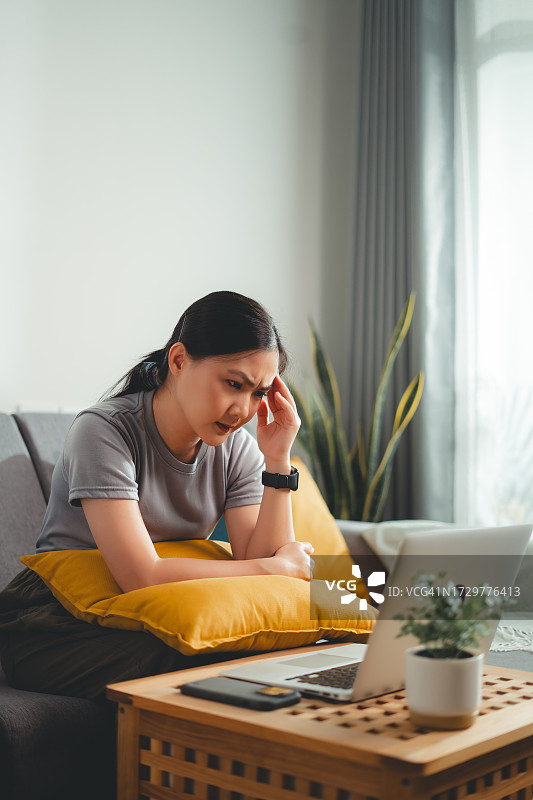 亚洲女人在家中客厅使用笔记本电脑，头痛难忍图片素材