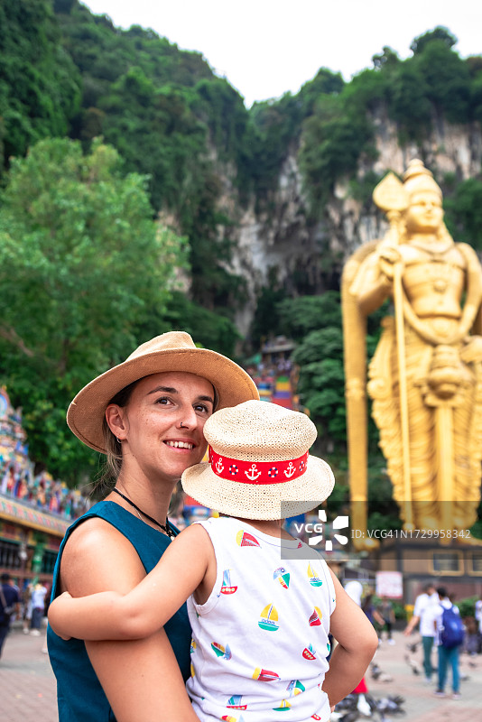 微笑女人抱着孩子看着镜头，背景是马来西亚的黑风洞图片素材