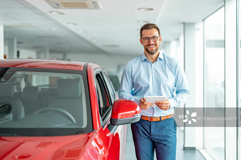 在豪华车沙龙站在新车旁，拿着平板电脑的快乐销售经理图片素材