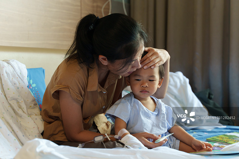 生病的亚洲儿童在医院接受母亲的照顾，使用体温计检查体温，接受抗生素药物和输液，等待康复。婴儿保健医疗概念图片素材