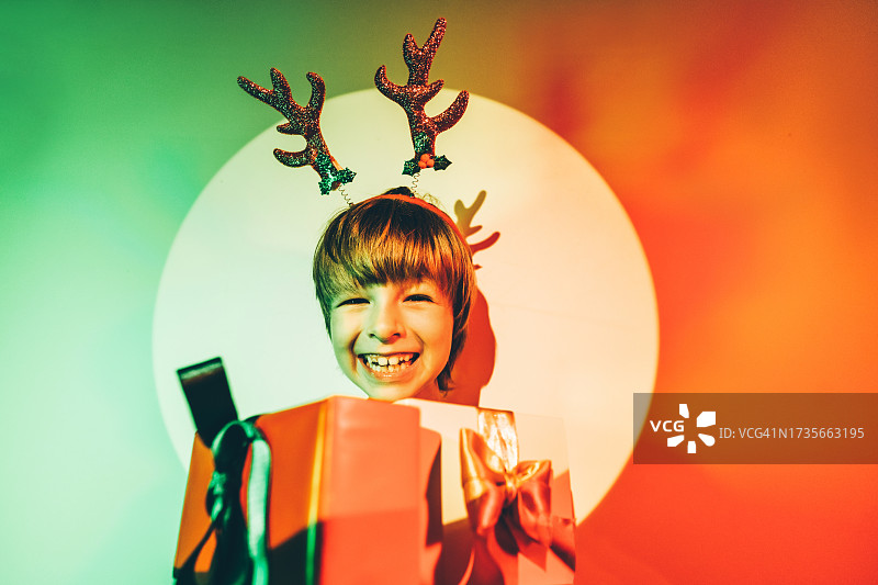 戴着驯鹿角并在圣诞节期间拿着圣诞礼物的有趣男孩。图片素材