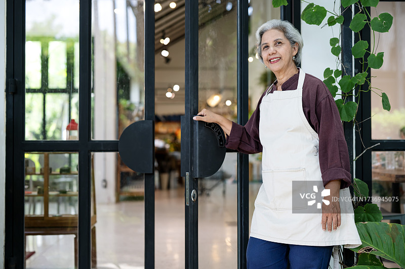 我们随时为您服务。每份菜单都保证质量。一位亚洲老年女性店主在她开业的餐厅前为顾客服务。本地餐饮业务概念。图片素材