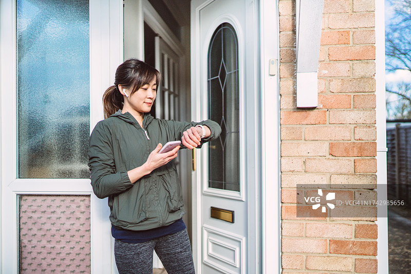 年轻漂亮的亚洲女性在开始在附近跑步前，将她的智能手表和智能手机上的运动追踪应用程序连接起来图片素材