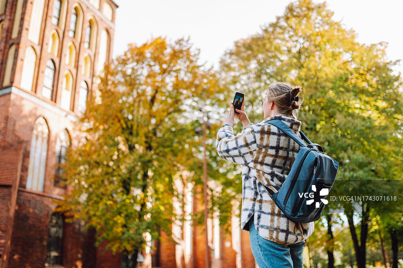城市秋日探险：金发女游客在阳光明媚的日子里拍摄公园绿地的地标图片素材