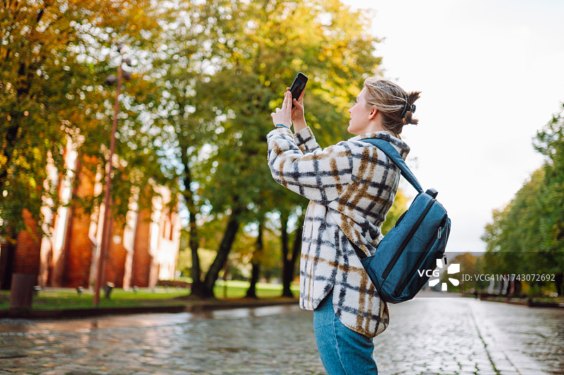 在秋季户外城市用智能手机拍照的背背包白人金发女子图片素材