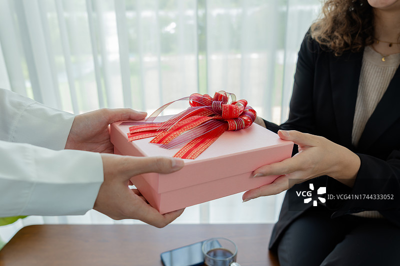 卷发的女人穿着工装，在阳光明媚的办公室里将圣诞礼物送给穿着白衬衫的同事，两位同事互相交流幸福，以加强员工敬业度和工作场所关系图片素材