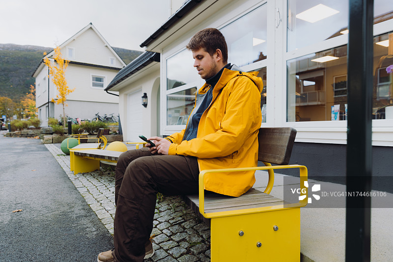 在城市街道长椅上使用智能手机的黄色夹克男人图片素材