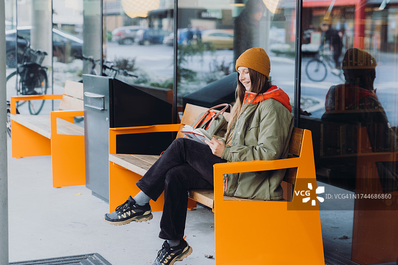 微笑的女人坐在现代城市街道的橙色长椅上看书图片素材