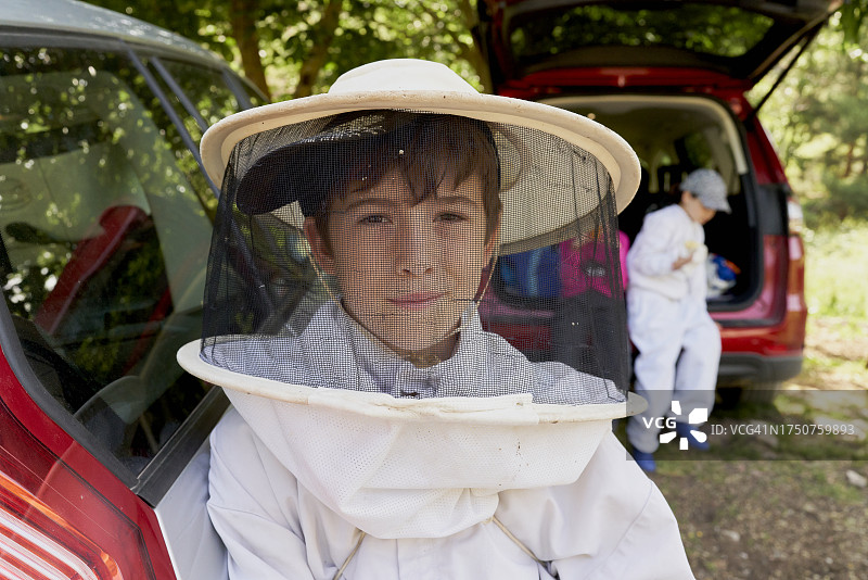 穿着养蜂防护服坐在汽车后备箱上对着镜头微笑的男孩图片素材