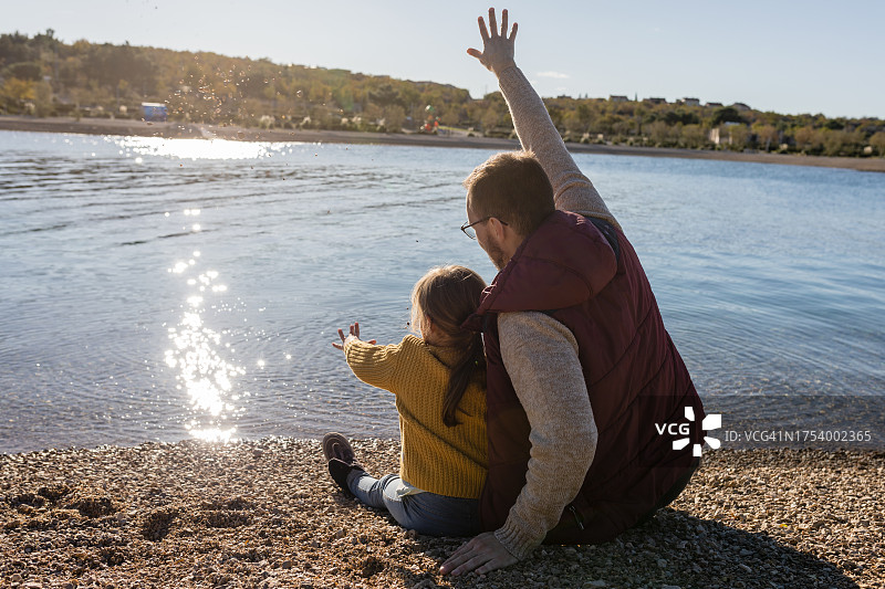父女坐在海滩上，从背后看，向水中扔石头图片素材