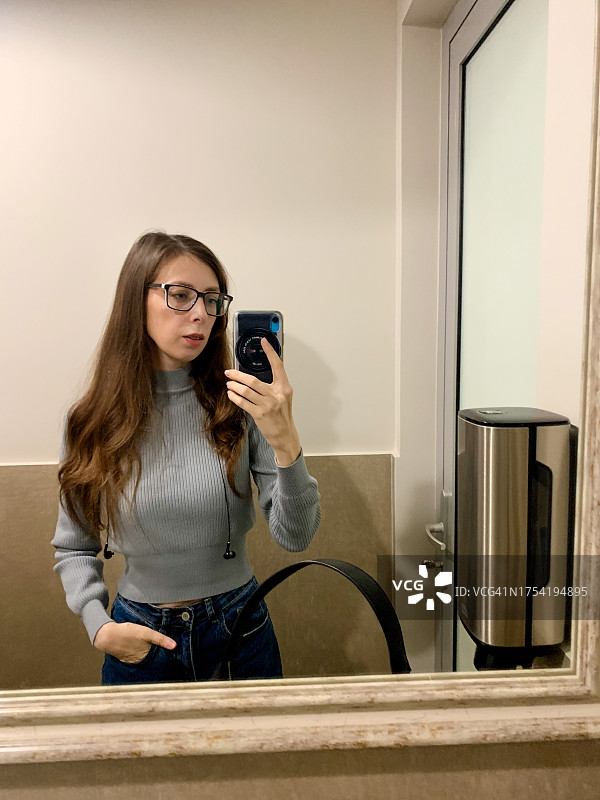 戴眼镜的长发年轻女人在咖啡馆的卫生间镜子前自拍，穿着灰色针织毛衣图片素材