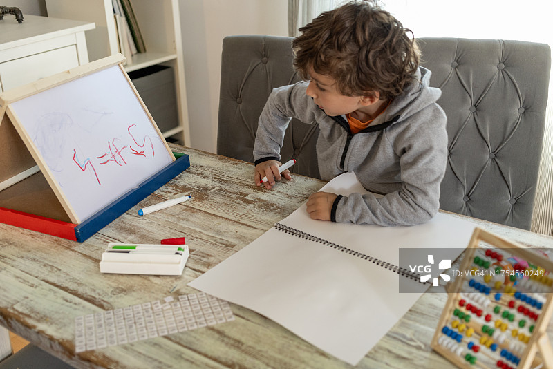 七岁卷发男孩在客厅餐桌旁认真学习功课,学龄前弟弟妹妹在一旁观看图片素材