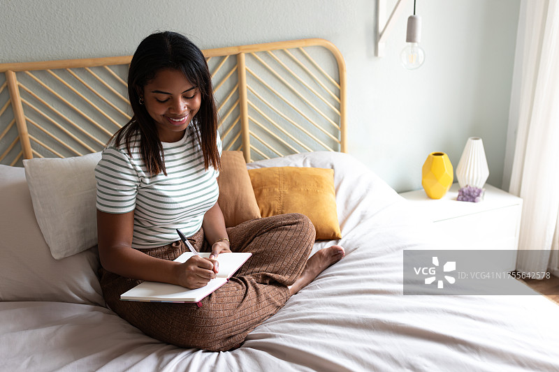 舒适卧室：快乐的年轻非裔美国妇女坐在床上写日记图片素材