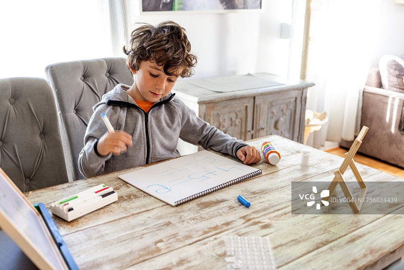 七岁卷发男孩在客厅餐桌旁认真学习功课,学龄前弟弟妹妹在一旁观看图片素材