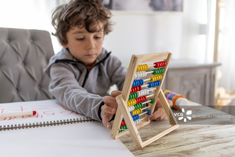 七岁卷发男孩在客厅餐桌旁认真学习功课,学龄前弟弟妹妹在一旁观看图片素材