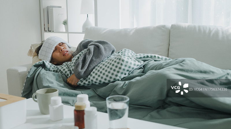 生病的亚洲年轻女性头痛发烧咳嗽感冒打喷嚏，躺在客厅沙发上的毯子下。医疗保健，居家隔离，保持社交距离。图片素材
