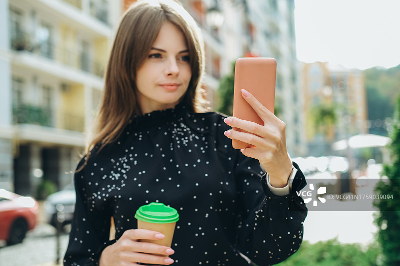 年轻女子靠在建筑物旁用纸杯喝水和使用智能手机图片素材