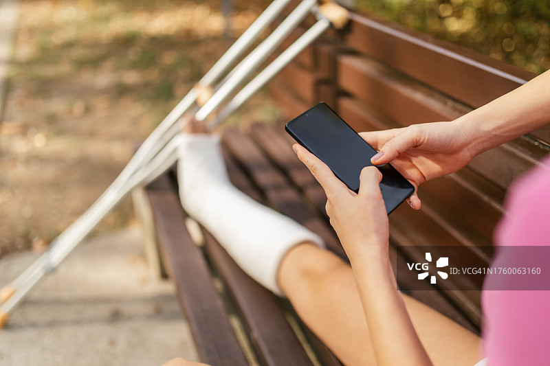 坐在公园长椅上、腿上打着石膏、使用手机的女人图片素材