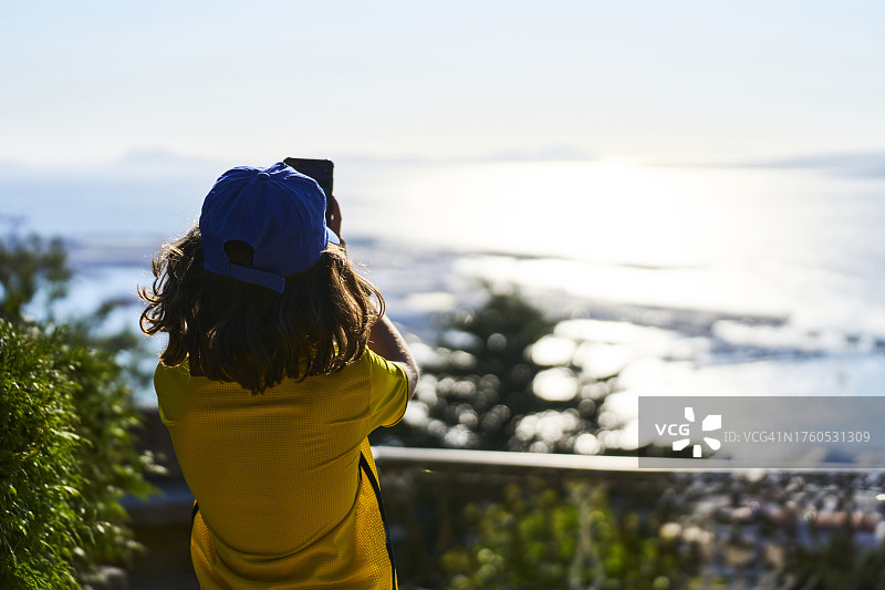 一个戴着帽子，穿着休闲装的女孩，用数码相机拍摄海岸的背影图片素材