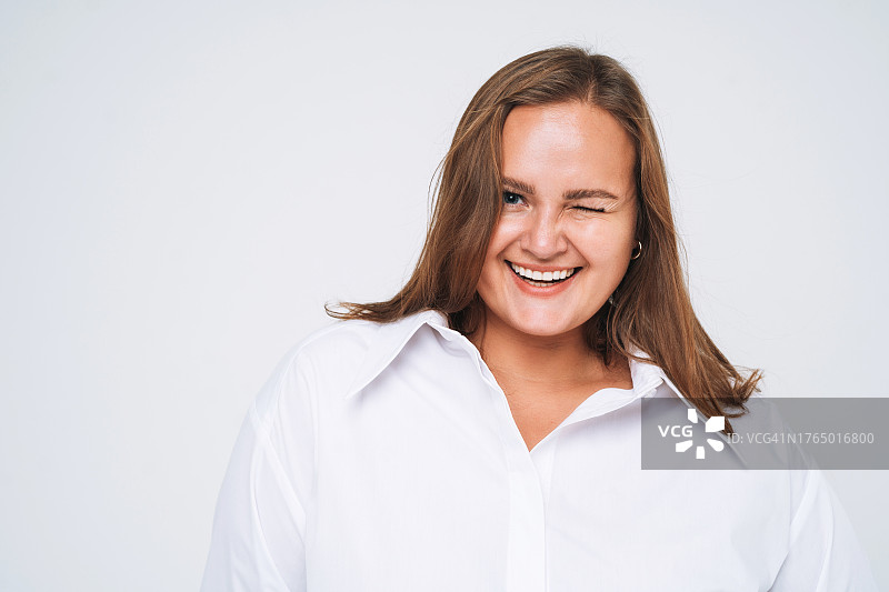在白色背景下眨眼的穿着白色衬衫的微笑的加大码年轻黑发女子肖像图片素材