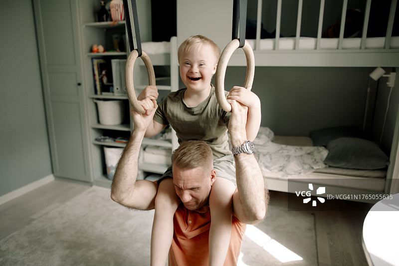 父亲扛着患有唐氏综合征的儿子，同时在卧室里拿着体操吊环图片素材