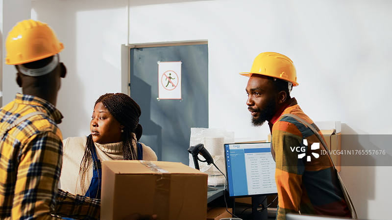 仓库工人佩戴安全帽，在办公桌旁手持包裹，使用电脑进行讨论图片素材