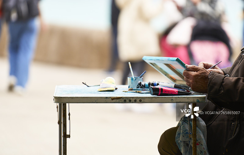 穿着外套的男人坐在户外桌旁在木板上绘画的侧视图图片素材