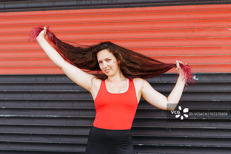一个穿着健身服的迷人运动型女孩拿着她的头发。一个穿着红色上衣和黑色紧身裤，留着长长的卷曲突出头发的迷人年轻女人微笑并玩得很开心。图片素材