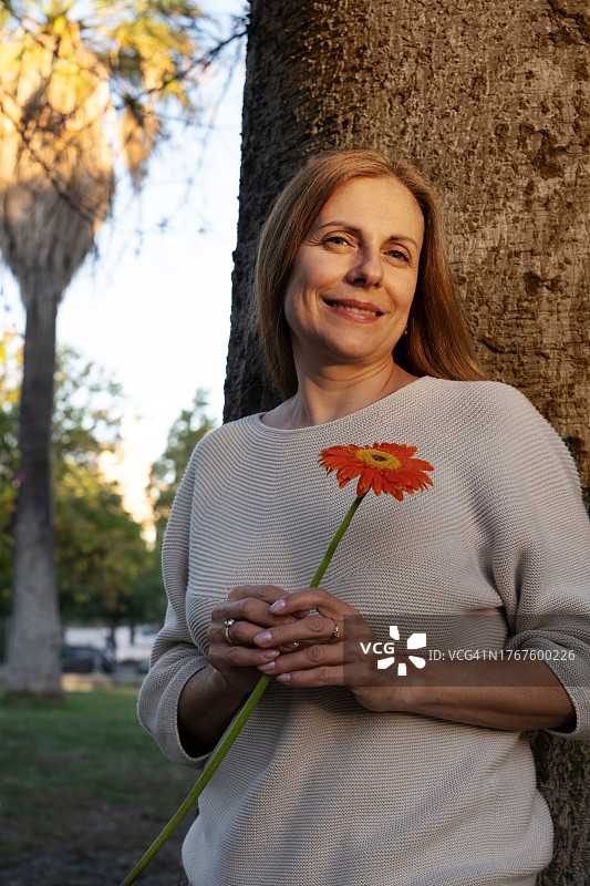富有魅力的成熟女人，留着红色长发，手持鲜花，站在公园的树旁，在城市街道上图片素材
