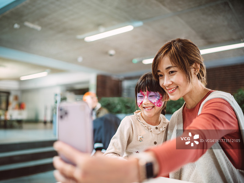 在主题公园咖啡馆，快乐的女孩和妈妈用手机自拍，脸上画着美丽的彩绘图片素材