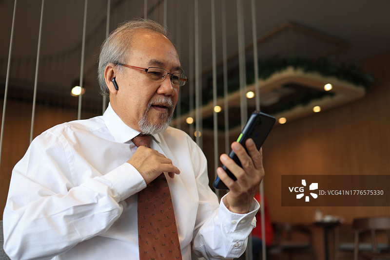 一位笑容满面的灰发年长男商人戴着无线耳机，用智能手机打电话图片素材