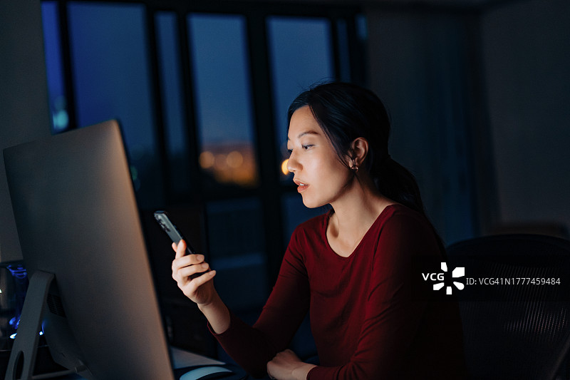 专注的年轻亚洲女性坐在电脑前，一边看手机一边工作，在家工作到很晚。在家办公。自由职业者，自主创业。在线学习。技术工作概念图片素材