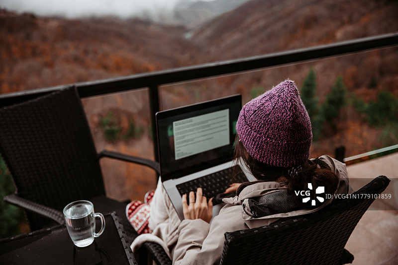 一位年轻女子在山间小屋远程办公，欣赏着壮丽的景色，使用笔记本电脑图片素材