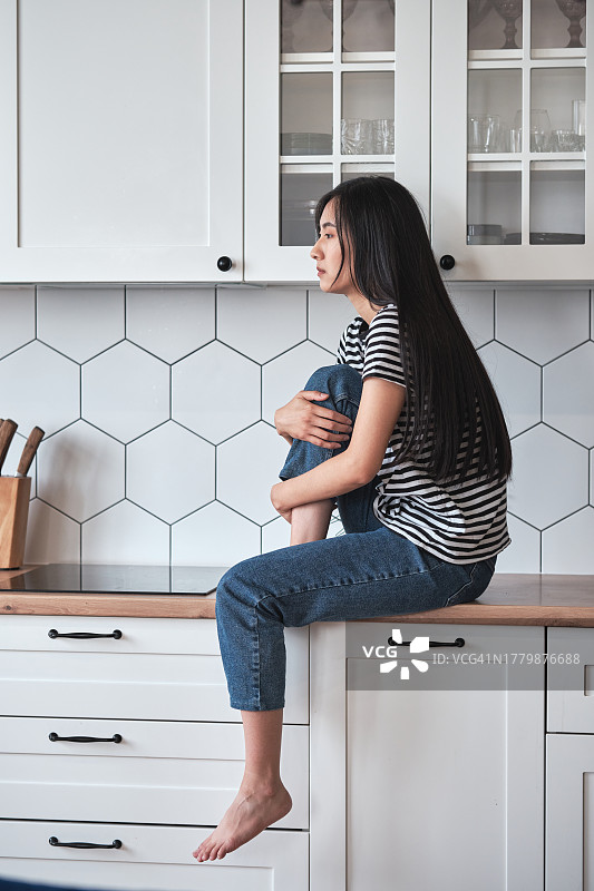 悲伤的亚洲女人坐在厨房的台面上，拥抱着膝盖，望着窗外，感到沮丧和孤独图片素材