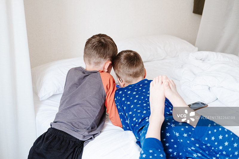 躺在床上玩手机的两个男孩和兄弟，戴着助听器的弟弟看着哥哥玩手机图片素材