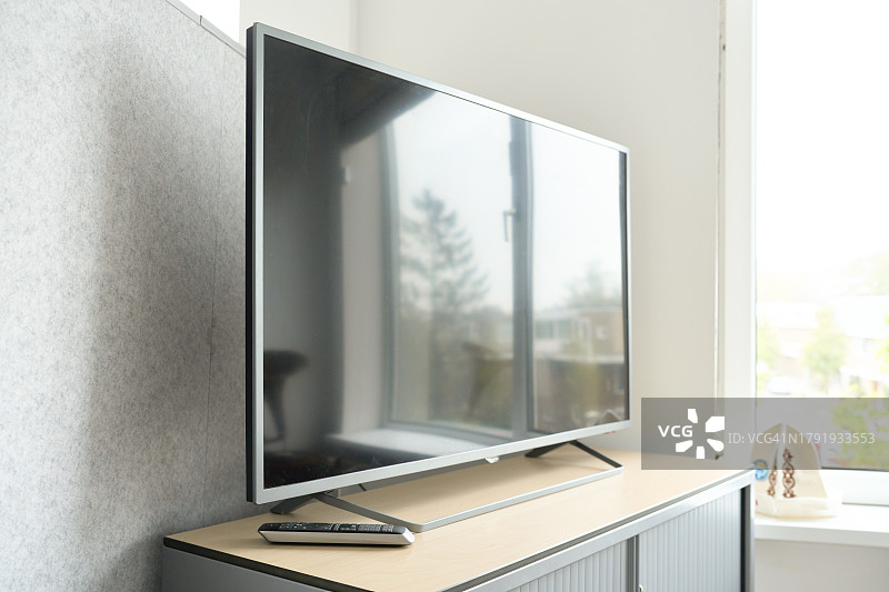 大屏幕电视关闭，屏幕反射窗户影像，遥控器置于桌面，背景为灰色墙壁图片素材