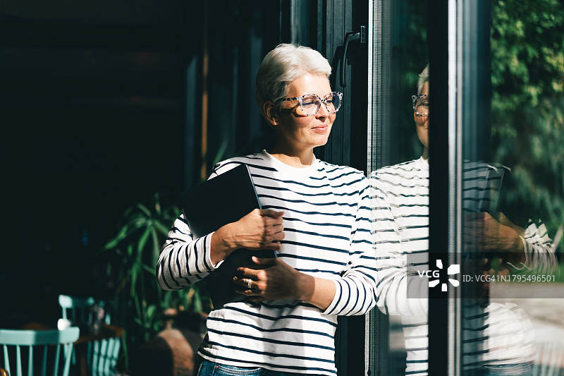 一位戴着眼镜、留着灰色短发的中年妇女在咖啡馆用笔记本电脑工作。企业主图片素材