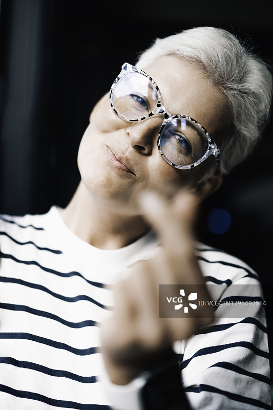 时尚的银色短发女子戴着图案眼镜，自信地微笑，并以顽皮的态度向镜头展示手指组成的心形符号图片素材