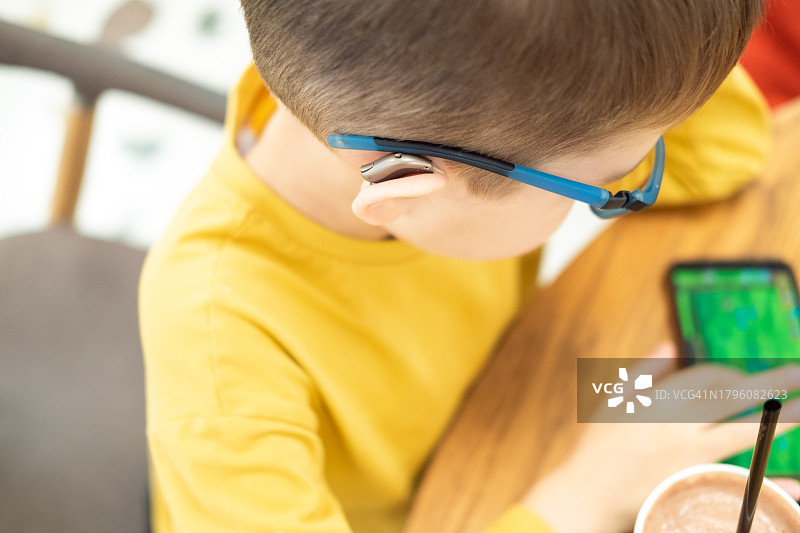 9岁的金发男孩戴着眼镜和黄色T恤坐在咖啡馆里，戴着助听器玩手机，顶视图图片素材
