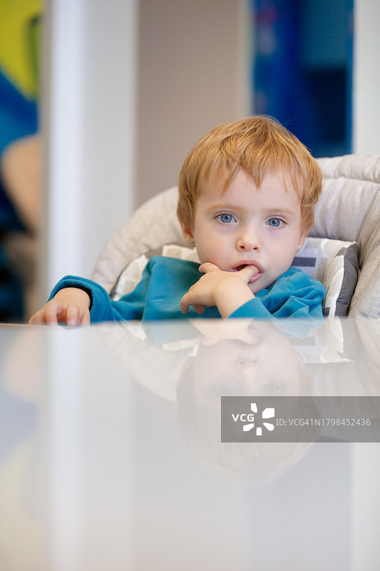 坐在高脚椅上，吮吸着拇指看着镜头的小金发男孩图片素材