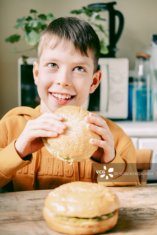11岁男孩在家里的厨房的桌旁，穿着橙色连帽衫咬着一个大肉饼汉堡图片素材