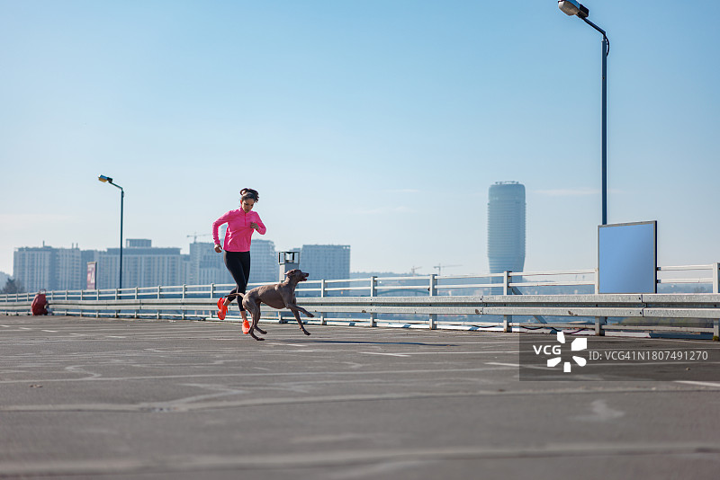 在一个晴朗的早晨，活力年轻女人在空旷的停车场顶上和她的狗一起慢跑图片素材