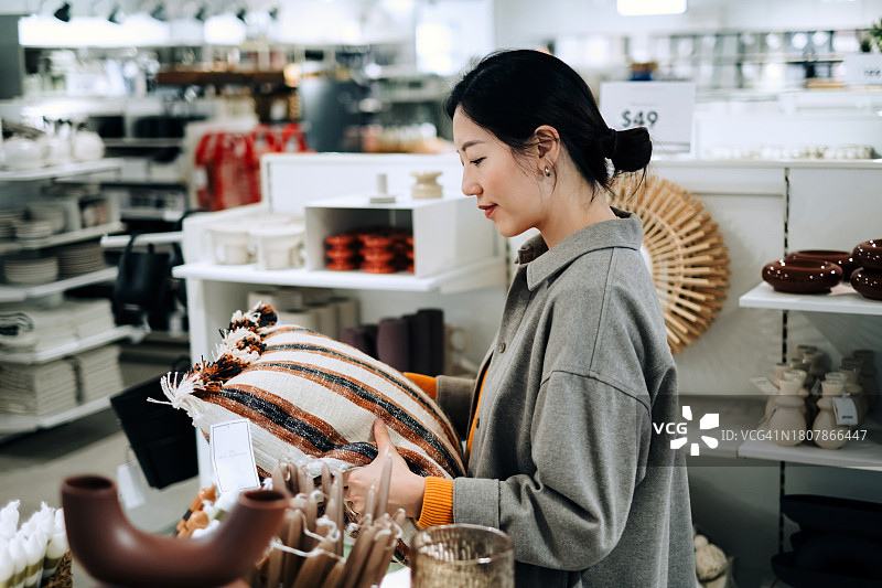 年轻亚裔女性在生活家居店选购家居装饰品，正在看一些展示的靠垫。生活方式、零售和购物概念图片素材