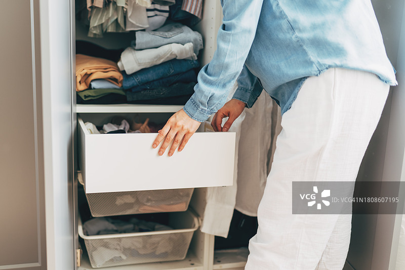 快乐的年轻红发女子在衣柜清洁时将衣服和内衣放入储物箱中。微笑的女性整齐地将衣服放入橱柜现代存储系统中，舒适的整理图片素材