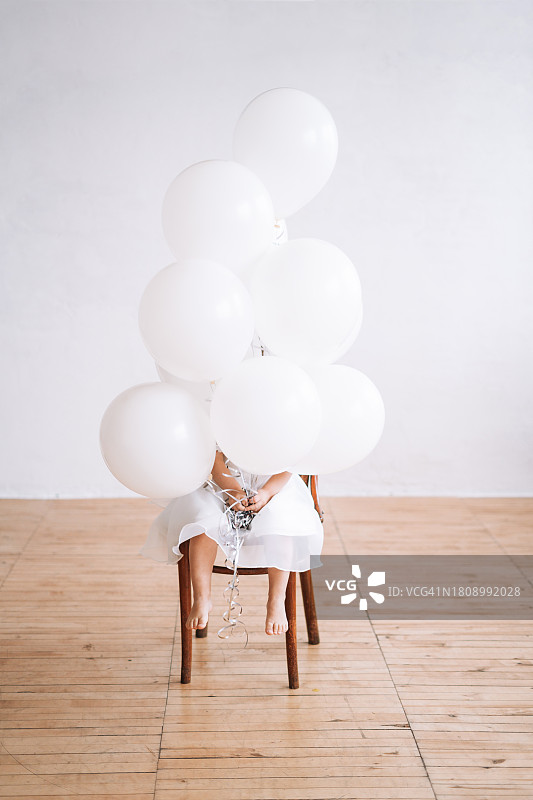 两岁可爱小女孩在明亮房间的儿童派对上穿着节日白色连衣裙，手拿充气气球图片素材